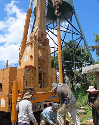 Cuatro trabajadores operan una maquinaria amarilla de perforación junto a la torre de un tanque de agua