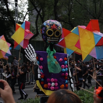 Una figura de calavera vestida de morado junto a personas que llevan estrellas de papel de colores durante un desfile en la vía pública