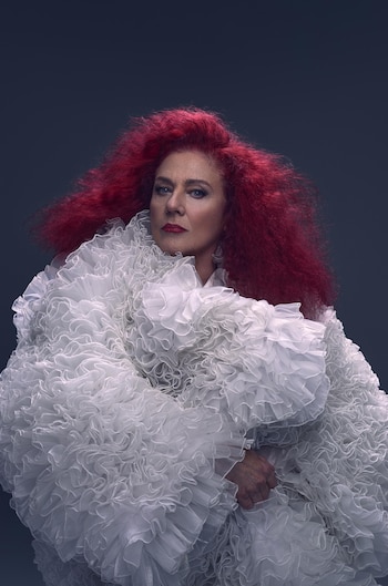 Una mujer con amplio cabello rojo vestida con una prenda de volantes blancos posa ante un fondo oscuro