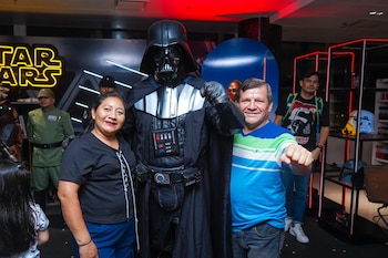 Una mujer y un hombre sonríen mientras posan junto a una persona vestida con un disfraz de Darth Vader. Otros fans y objetos de Star Wars se ven al fondo