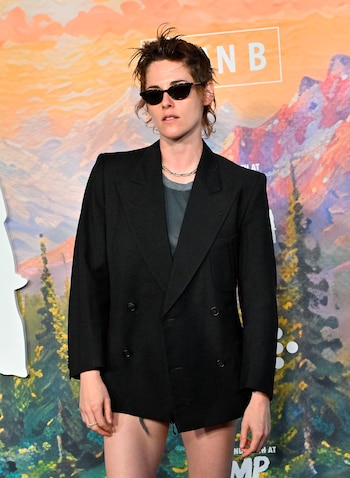 Kristen Stewart con pelo corto desordenado, gafas de sol, chaqueta negra, camiseta gris, collar plateado y fondo montañoso