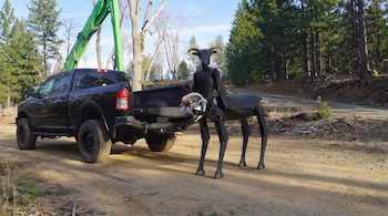 Robot cuadrúpedo negro con cuernos, motosierra, camioneta pickup negra, brazo de grúa verde, camino de tierra, árboles, cielo azul, troncos en el suelo.