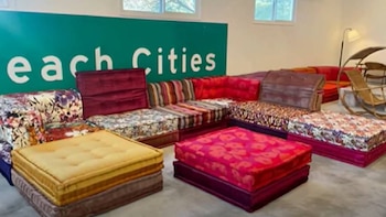 Sofás modulares, colchones y futones coloridos con variados estampados (floral, rayas) dispuestos como cojines de suelo. Suelo gris, pared verde con 'Cities'