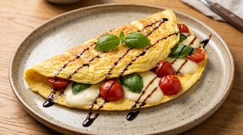 Omelette de huevo con relleno de tomates cherry, queso mozzarella, hojas de albahaca y salsa. Servido en un plato redondo sobre una mesa de madera.