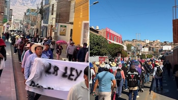 Una multitud de personas camina por una calle urbana; algunos manifestantes portan sombreros y uno lleva una pancarta. Edificios y montañas al fondo