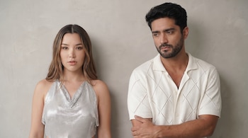 Retrato de una mujer con cabello castaño claro y un top plateado, de pie junto a un hombre con barba y camisa color crema, sobre un fondo gris claro.