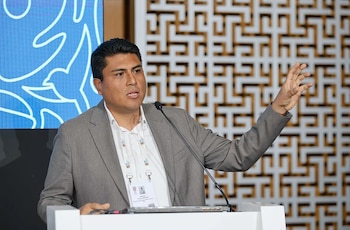 Hombre de traje gris y camisa blanca con credencial al cuello habla en podio con micrófono. Al fondo, pantalla azul con figura y pared con patrón geométrico