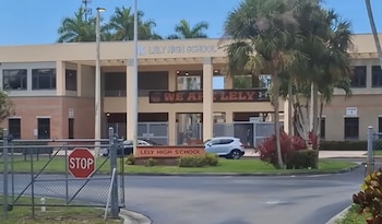Vista exterior de Lely High School con su nombre en la fachada y un cartel en el césped, rodeada de palmeras y vehículos, bajo un cielo claro