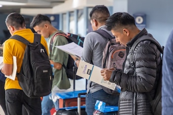 Vista lateral de cinco jóvenes con mochilas y chaquetas. Sostienen y leen documentos en un interior, un hombre al frente lleva chaqueta acolchada