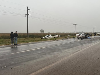 Vista de una carretera mojada con dos vehículos accidentados: una camioneta blanca en la zanja y un sedán blanco con daños graves, con escombros y personas observando