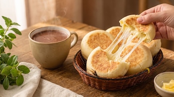 Una mano sujeta un pan de queso con hebras de queso fundido que se estiran desde una canasta, junto a una taza humeante y trozos de mantequilla.