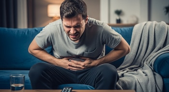 Un hombre con camiseta gris y pantalones oscuros está sentado en un sofá azul sujetándose el abdomen. Una manta gris, un vaso de agua y un mando a distancia se ven en la imagen.