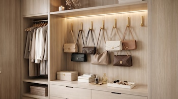 Un armario de madera clara muestra ropa colgada, siete bolsos de diseño en ganchos dorados, y accesorios como gafas de sol y cajas sobre cajones.