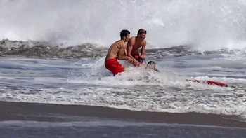 Dos hombres en el agua, uno con bañador rojo y otro con bañador estampado, ayudan a una persona parcialmente sumergida. Hay olas, espuma y un flotador de rescate rojo