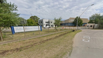 Un cartel con la leyenda Adler Pelzer Group frente a un predio industrial cercado con edificios, árboles y un camino pavimentado