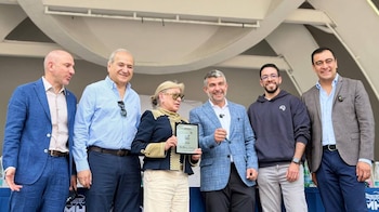 Un grupo de cinco hombres y una mujer posa al aire libre en un evento; la mujer al centro sostiene un reconocimiento junto a Mauricio Tabe, con fondo arquitectónico