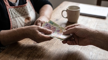 Manos de un empleador que entregan billetes de pesos argentinos a una mujer con delantal junto a una taza sobre una mesa.