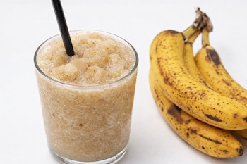 Un vaso transparente con un granizado de plátano y una pajita negra. Al lado derecho, un racimo de plátanos maduros con manchas oscuras.