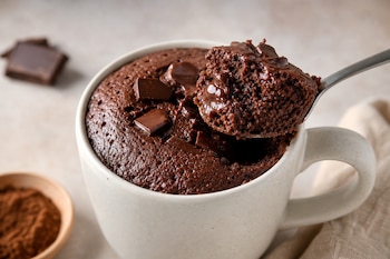 Taza de cerámica con bizcocho de chocolate, una cuchara que sostiene un bocado, trozos de chocolate y un cuenco con cacao en polvo.