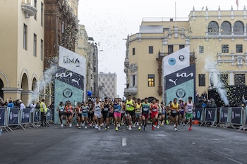Numerosos corredores en una calle urbana, con edificios de fondo. Hay dos carteles de inicio de carrera, confeti en el aire y vallas de seguridad