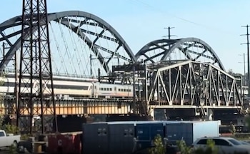 Un tren de pasajeros cruza una estructura ferroviaria junto a arcos metálicos de puente, torres de electricidad y contenedores