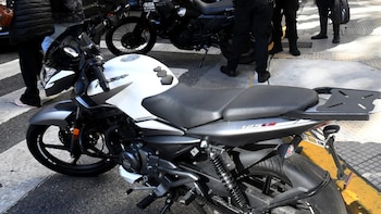 Primer plano de una motocicleta blanca y gris estacionada. Detrás se ven otras motocicletas oscuras, una con la inscripción "POLICIA DE LA CIUDAD", y agentes uniformados