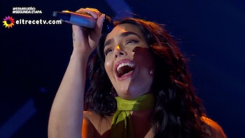 Primer plano de una cantante de cabello oscuro y pañuelo verde, con la boca abierta y el micrófono en alto, actuando en un escenario oscuro con luces azules