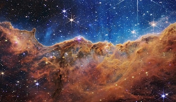 Nubes anaranjadas y marrones de polvo y gas se extienden en la parte inferior frente a un fondo azul lleno de estrellas brillantes