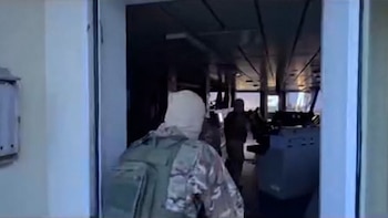 Vista trasera de un soldado con uniforme de camuflaje y pasamontañas claro entrando a un puente de mando oscuro de un barco, con otra figura al fondo