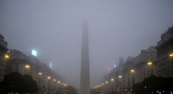 Vista frontal del Obelisco de Buenos Aires cubierto por una densa niebla nocturna, rodeado por edificios urbanos y farolas encendidas a los lados.
