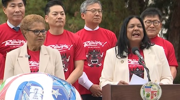 Un grupo de personas, incluyendo dos mujeres con chaquetas claras y varios hombres con camisetas rojas, están en un podio con un micrófono y un balón de la Copa Mundial