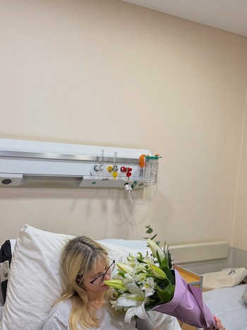Denisse Gonzalez, una mujer rubia con gafas, recostada en una cama de clínica, sostiene un ramo de flores blancas envuelto en papel morado