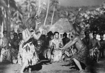 Fotografía en blanco y negro con dos bailarines de tocados y faldas tradicionales. Al fondo, espectadores, palmeras y una cabaña de paja