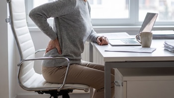 Persona sentada en una silla de oficina blanca. Su mano derecha está en la espalda baja. Un portátil, una taza y papeles sobre el escritorio. Ventana de fondo