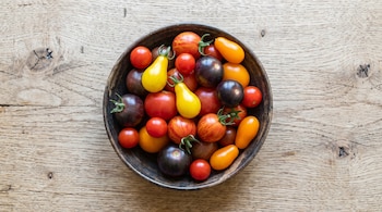 Un cuenco oscuro con tomates de varios colores: rojo, amarillo, anaranjado, morado oscuro y rayados. La mesa de madera clara tiene vetas visibles.