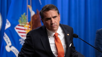 Retrato de Todd Blanche, un hombre caucásico de mediana edad con cabello oscuro, vistiendo un traje oscuro y corbata naranja. Está frente a una bandera azul con insignias