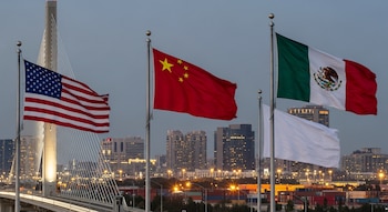 Cuatro mástiles con las banderas de Estados Unidos, China, México y una bandera blanca, un puente atirantado, un horizonte urbano y contenedores de carga.