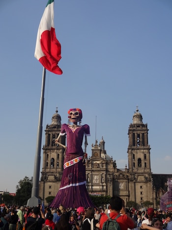 Una figura gigante de la Catrina con vestido morado, una bandera de México ondeando en un asta, la Catedral Metropolitana y una multitud reunida