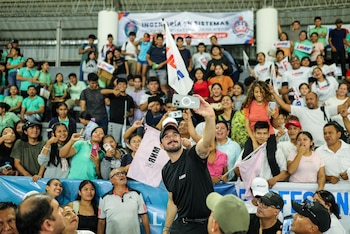 Juan Pablo Velasco, con gorra oscura y camiseta negra, sonríe mientras se toma una selfie con un celular plateado, rodeado de una multitud de personas diversas