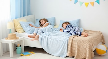 Niña y niño duermen en una cama con sábanas azules, junto a una mesa con lámpara y sombrero de paja, y una pelota de playa en el suelo.