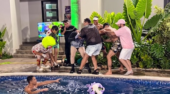 Nueve hombres junto a una piscina, algunos en una interacción física, uno en el agua, uno con disfraz de payaso, plantas y una televisión encendida.