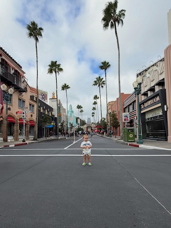 Un niño pequeño de pie en el centro de una calle asfaltada flanqueada por palmeras altas y fachadas de edificios de estilo vintage