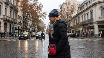 Mujer con campera oscura, gorro de lana y chalina a rayas cruzando una calle mojada de adoquines en Buenos Aires, con coches y edificios de fondo.