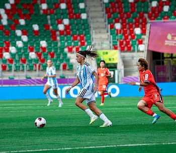 Una jugadora con la camiseta a rayas celestes y blancas corre tras el balón frente a una rival vestida de rojo