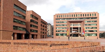 Edificios de ladrillo rojo rodeando una plaza con peatones, banderas en astas y un muro bajo en primer plano
