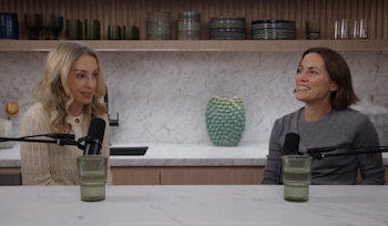 Dos mujeres sentadas frente a micrófonos y vasos, con estantes con vajilla y una pared de mármol al fondo