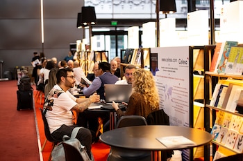 Personas interactúan en un concurrido stand de libros. Se ven mesas redondas, un hombre y una mujer con un portátil, estanterías con libros y un cartel de Celesa