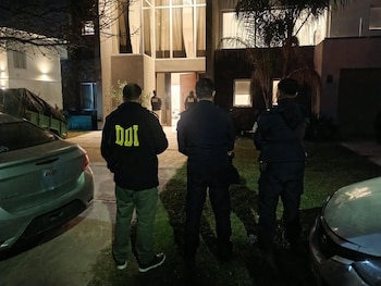 Tres efectivos policiales de espalda, uno con las siglas DDI en su chamarra, observan una vivienda iluminada durante la noche