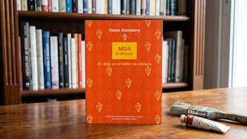 Primer plano de la portada naranja del libro 'MDA El Manual' de Diana Aisenberg sobre una mesa de madera; pinceles y un tubo de pintura a la derecha, estanterías al fondo