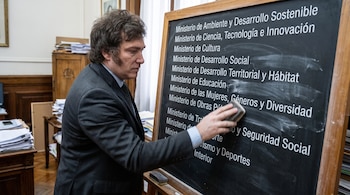 Javier Milei sostiene un borrador mientras limpia una lista de ministerios escrita en blanco sobre un pizarrón de madera.